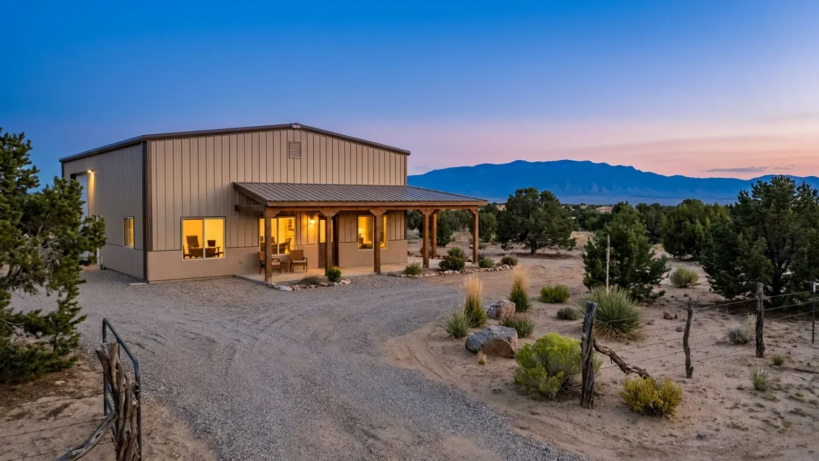 A finished tan barndominium at dusk with warm interior lighting glowing through its large windows and a mountain range behind.