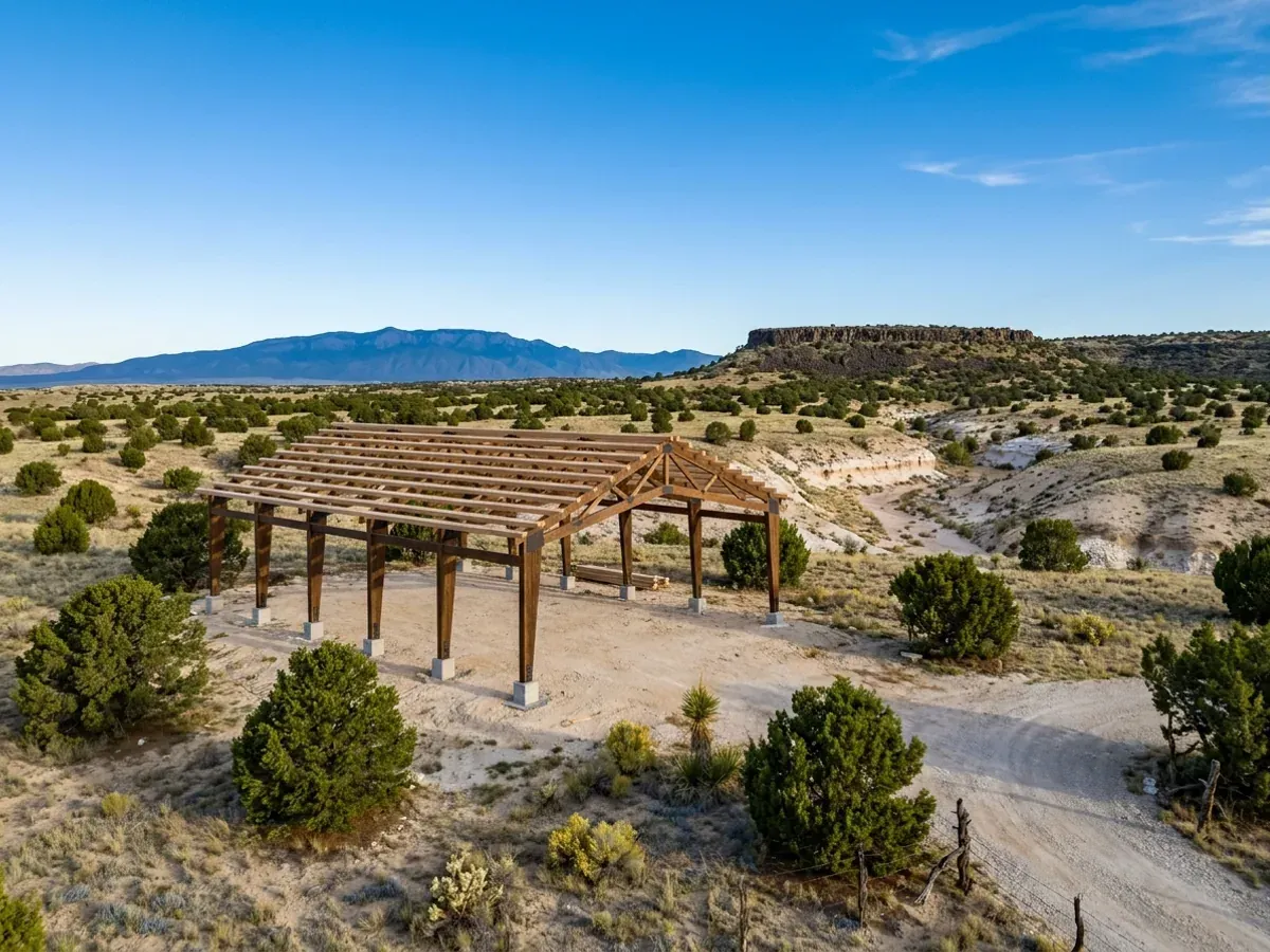 A post-frame building under construction in open high desert, heavy timber columns carrying the roof framing, with a mesa and mountains beyond.