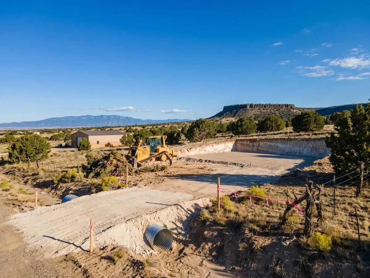 A dozer clearing brush beside a graded building pad on a high desert parcel, with pale caliche in the cut face and a culvert set at the driveway.